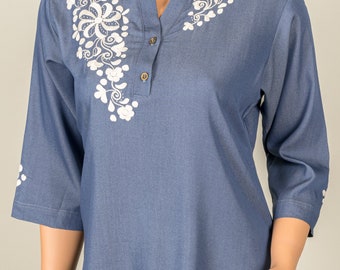 May include: A blue denim shirt with white embroidered floral designs on the shoulders. The shirt has a mandarin collar and a single button closure.