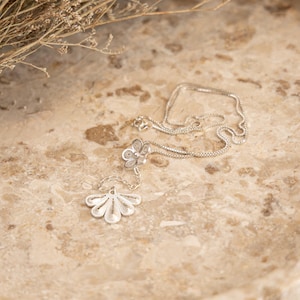 May include: A delicate silver necklace with a floral pendant and a fan-shaped charm. The necklace is displayed on a light brown, marble-like surface, with dried plants in the background. The pendant and charm have intricate filigree details.