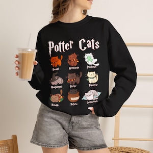 Könnte beinhalten: Ein schwarzes Langarm-Sweatshirt mit dem Schriftzug Potter Cats in weißen Buchstaben über neun illustrierten Katzen. Jede Katze ist mit einem Namen beschriftet, darunter Rawnald, Hermeownle, Pawdamon, Meowgonagall, Purrter, Meowfoy, Nyaville, Sefurus und Dumpurrdore.