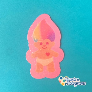 May include: A pink sticker featuring a baby troll doll with a rainbow-colored hair, a red heart, and a white diaper. The sticker has a glittery outline and the text "Hearts n Holograms" in the bottom right corner.