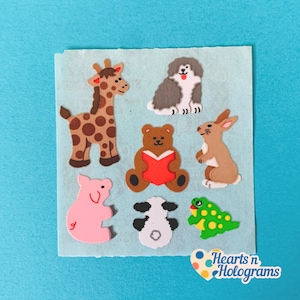May include: A collection of felt animal stickers, including a giraffe, dog, bear, rabbit, pig, cow, and frog. The giraffe is brown with dark brown spots. The bear is reading a red book. The stickers are on a light blue background.