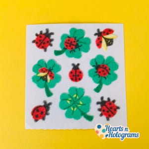 May include: A sheet of stickers featuring red ladybugs and green shamrocks. The ladybugs have black spots and are set against a white background. The stickers are on a yellow background. The text "Hearts n Holograms" is in the bottom right corner.
