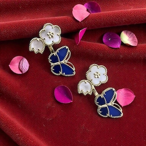 Pode incluir: Duas broches douradas em forma de flor e borboleta com detalhes em esmalte branco e azul. As broches estão colocadas sobre um fundo de veludo vermelho, com pétalas de flores rosa e roxas espalhadas.