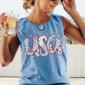 Floral USA Comfort Colors tank top in light blue, styled on a woman enjoying lemonade outdoors. This patriotic 4th of July tank for women features a red white and blue graphic, perfect for summer BBQ outfits, America shirts, or cute July Fourth looks.