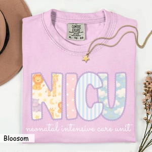 May include: A light purple t-shirt with the letters "NICU" in a decorative font, each letter featuring a different pattern and animal illustrations. The text "neonatal intensive care unit" is below the letters. A gold star necklace rests on the shirt.