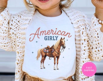 May include: White t-shirt featuring the text "American Girly" in red and blue script above an illustration of a brown horse with a saddle. The design includes small star accents.