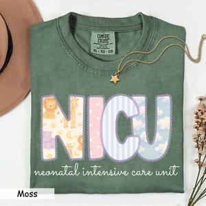 May include: Moss green t-shirt with the letters "NICU" in a whimsical design featuring animals, clouds, and stripes. The words "neonatal intensive care unit" are printed below the letters. A gold star necklace rests on the shirt.