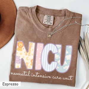 May include: Espresso-colored t-shirt with the letters "NICU" in pastel colors and animal patterns. The words "neonatal intensive care unit" are printed below. A silver necklace with a pendant is on the shirt.
