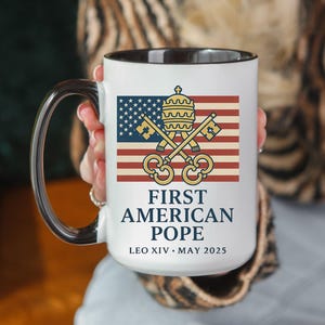 May include: White ceramic mug with a black interior and handle. The mug features a design with the American flag, crossed keys, and the text "FIRST AMERICAN POPE LEO XIV MAY 2025".