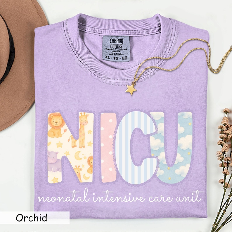 May include: Orchid-colored t-shirt with the letters "NICU" in a whimsical design featuring animals and patterns. The text "neonatal intensive care unit" is below the letters. A gold star pendant necklace rests on the shirt.