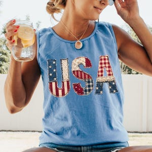 May include: Light blue tank top with the word "USA" appliqued on the front. The letters are made of various patriotic fabrics, including denim, gingham, and star-spangled patterns. The tank top is sleeveless and has a scoop neck.