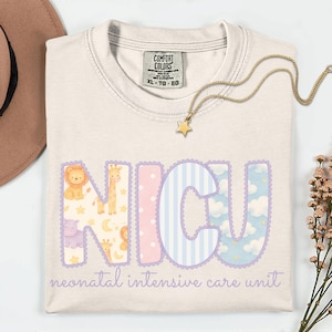 May include: Cream-colored t-shirt with the letters "NICU" in pastel colors and animal illustrations. The words "neonatal intensive care unit" are printed below. A gold necklace with a star pendant rests on the shirt.