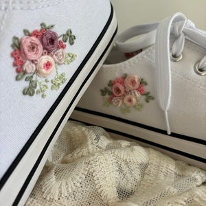 May include: A pair of white canvas shoes with pink and green embroidered floral designs. The shoes are sitting on a white lace fabric.