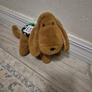 May include: A plush brown dog toy with floppy ears and a black nose. The toy has a black and white tag that says "ESS". The dog is sitting on a light-colored floor.