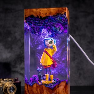 May include: A rectangular resin lamp featuring a miniature figure with blue hair in a yellow raincoat and a black cat. The clear resin contains swirling purple smoke effects, encased between two pieces of natural wood. A vintage camera sits nearby.