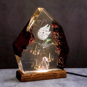 May include: A Star Wars-themed lamp with a clear resin body. The lamp features miniature models of the Millennium Falcon and X-wing fighters. The base is made of wood. The lamp is illuminated from within, creating a glowing effect, and is suitable for a desk or bedside table.