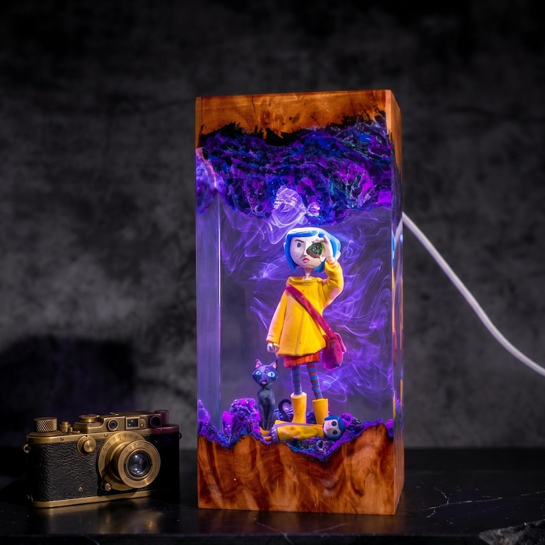 May include: A rectangular resin lamp featuring a miniature figure with blue hair in a yellow raincoat and a black cat. The clear resin contains swirling purple smoke effects, encased between two pieces of natural wood. A vintage camera sits nearby.
