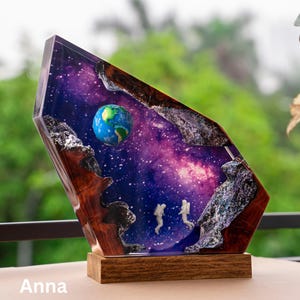 May include: A unique resin art piece featuring a miniature Earth and two astronaut figures set against a vibrant purple and blue galaxy backdrop. The artwork is encased in a geometric wooden frame with a wooden base. The name "Anna" is at the bottom.