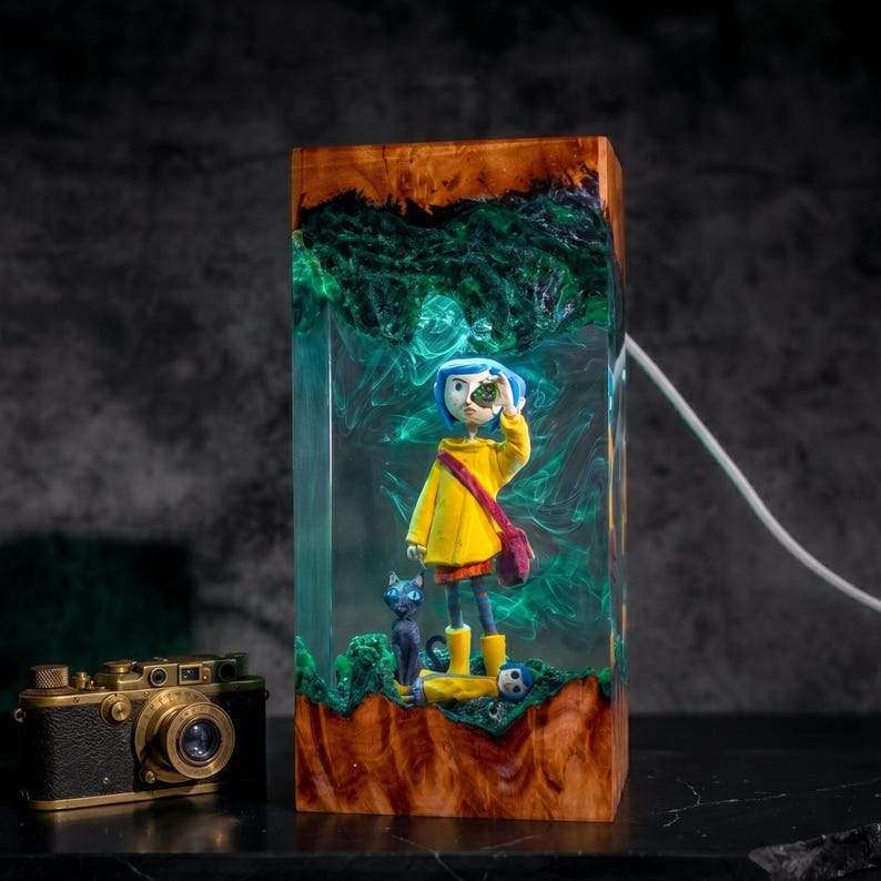 May include: A rectangular resin and wood lamp featuring a miniature figure with blue hair in a yellow raincoat holding a green object, accompanied by a black cat. The translucent green resin interior glows, encased between two polished wooden blocks.