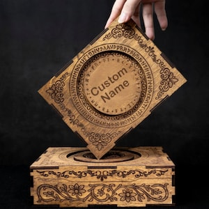 May include: A square wooden box with a removable lid featuring laser-engraved floral patterns and a circular center containing runic symbols and the text Custom Name. The box is crafted from light brown wood with dark brown engraved details.
