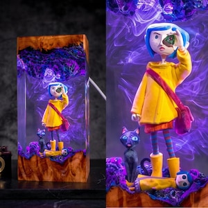May include: A rectangular resin and wood lamp featuring a miniature figure with blue hair in a yellow raincoat holding a green stone, accompanied by a black cat and a smaller figure in a yellow raincoat, set against a swirling purple smoke effect.