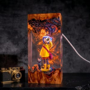 May include: A rectangular resin and wood lamp featuring a miniature figure with blue hair in a yellow raincoat, a black cat, and a small doll. The interior glows with orange light, and a white power cord extends from the side of the wooden base.