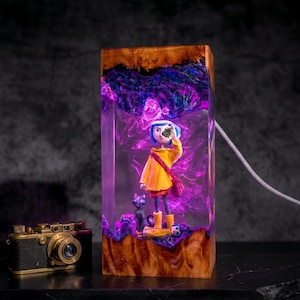May include: A rectangular resin and wood lamp featuring a Coraline figure in a yellow raincoat, a black cat, and a doll, illuminated by purple and blue light. Beside the lamp sits a vintage-style black and gold camera with a prominent lens.