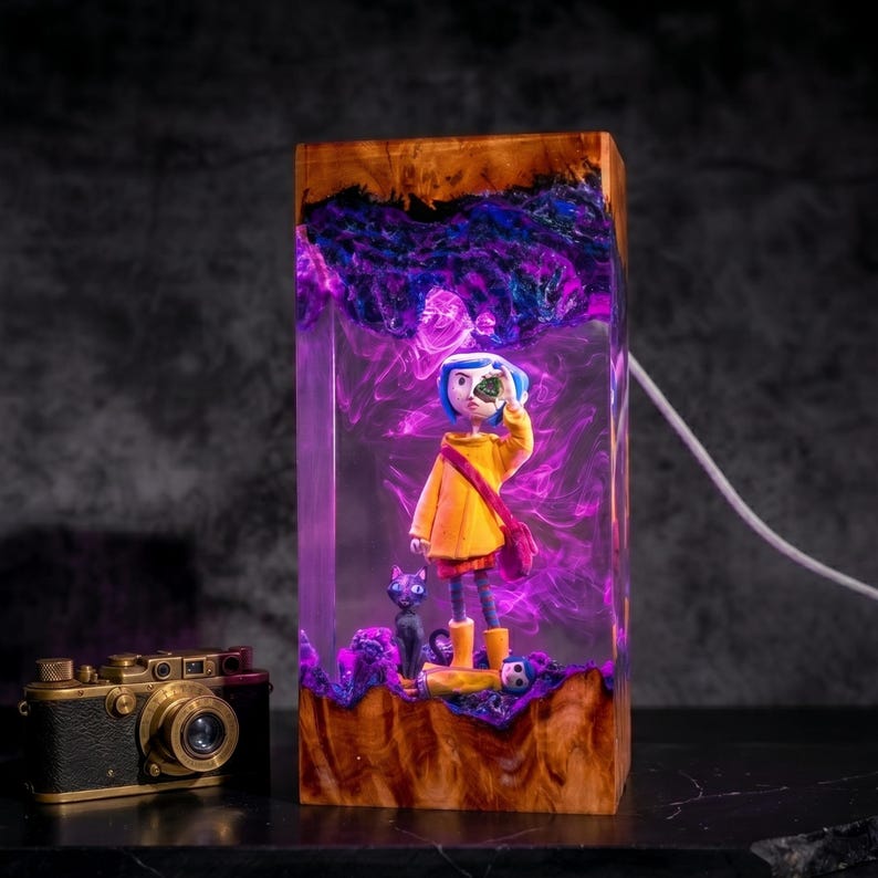 May include: A rectangular resin and wood lamp featuring a Coraline figure in a yellow raincoat, a black cat, and a doll, illuminated by purple and blue light. Beside the lamp sits a vintage-style black and gold camera with a prominent lens.