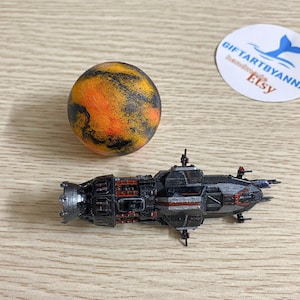 May include: A miniature black and silver spaceship model with red accents, next to a spherical planet model in shades of orange, yellow, and black. The Etsy shop logo is visible in the background.