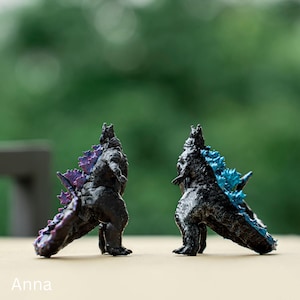 Könnte beinhalten: Zwei schwarze Godzilla-Figuren mit unterschiedlich gefärbten Stacheln. Eine Figur hat einen violetten Stachel und die andere einen blauen Stachel. Die Figuren sind in Kampfstellung.