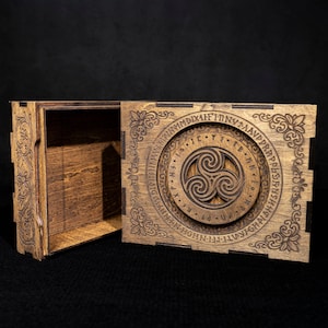 May include: A rectangular wooden box with a sliding lid featuring an intricate laser-engraved triskelion symbol surrounded by a circular band of runic-style characters and floral corner patterns. The box has a dark brown stained finish.