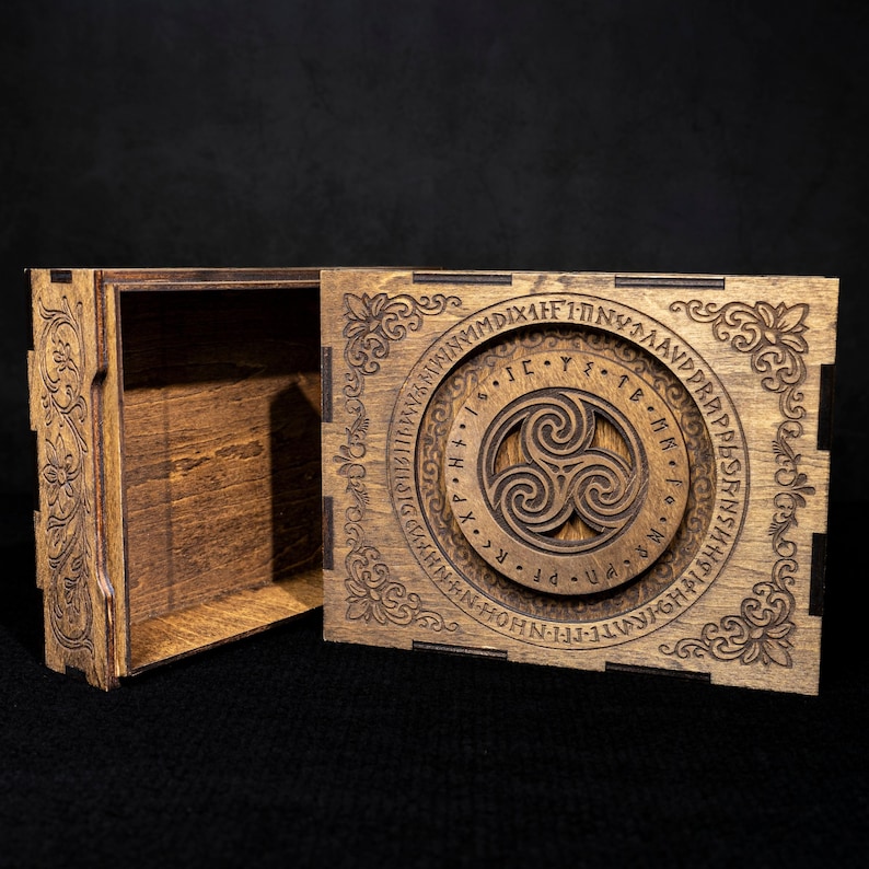 May include: A rectangular wooden box with a sliding lid featuring an intricate laser-engraved triskelion symbol surrounded by a circular band of runic-style characters and floral corner patterns. The box has a dark brown stained finish.