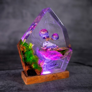 May include: A decorative resin lamp featuring a miniature landscape with a small creature, trees, and moss. The lamp has a wooden base and a clear, geometric resin structure with purple and pink hues. The lamp is lit from within, creating a glowing effect.