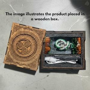 May include: A wooden box with a hinged lid featuring a circular Celtic triskele design and runic inscriptions. Inside, a rectangular resin block contains a coiled green dragon figure, accompanied by a white USB charging cable.