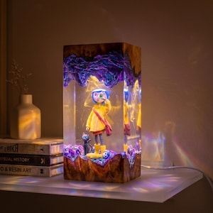 May include: A rectangular resin lamp featuring a figure with blue hair in a yellow raincoat and a black cat encased within clear resin. The top and bottom sections are made of dark wood with purple accents, and the lamp emits a warm internal glow.