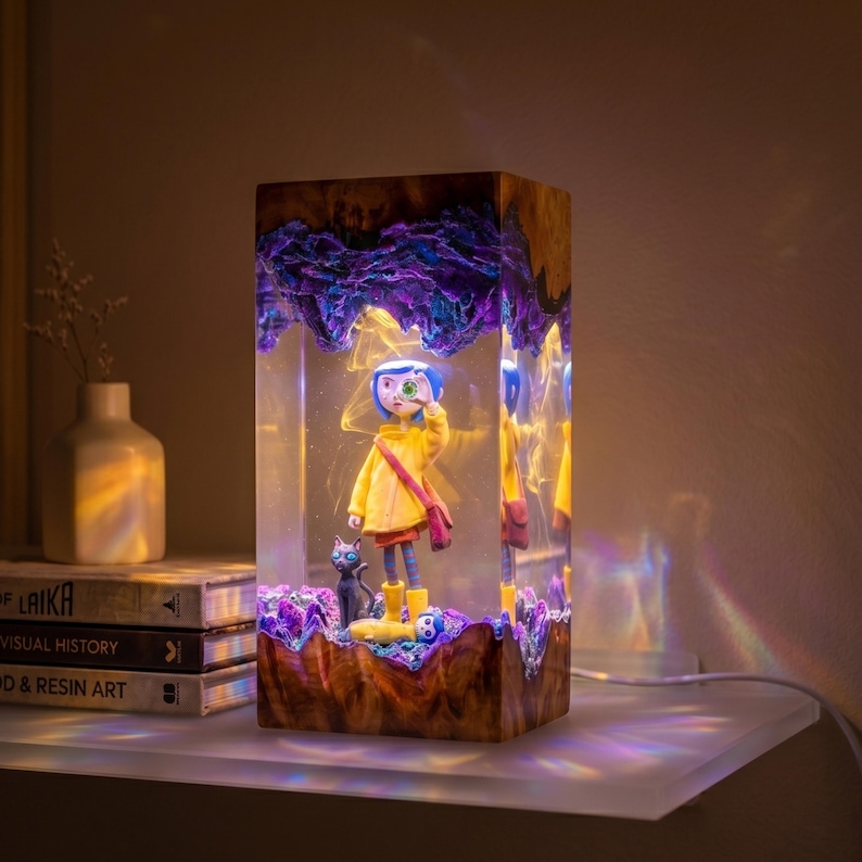 May include: A rectangular resin lamp featuring a figure with blue hair in a yellow raincoat and a black cat encased within clear resin. The top and bottom sections are made of dark wood with purple accents, and the lamp emits a warm internal glow.