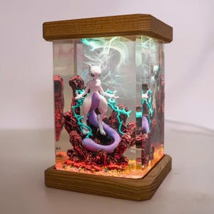 May include: A rectangular lamp featuring a Mewtwo figure inside a clear acrylic case. The figure is surrounded by red and purple rock formations and teal energy effects. The lamp has a wooden base and top.