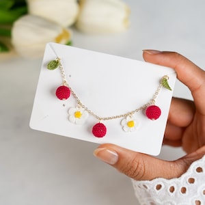 May include: A gold chain necklace featuring crochet charms. The necklace has red cherry and white daisy charms with yellow centers. Green leaves accent the chain. The necklace is displayed on a white card.