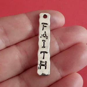 May include: Silver charm with the word "FAITH" embossed on it. The charm has a textured surface and a hole at the top for attaching to a necklace or bracelet.