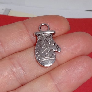 May include: Silver charm in the shape of a mitten with a textured design. The charm has a loop at the top for attaching to a bracelet or necklace.