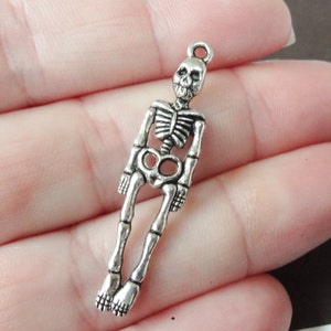 May include: A silver-colored skeleton charm with a loop at the top for hanging. The charm features detailed skeletal features, including a rib cage, skull, and leg bones. The charm is approximately 2 inches in length.