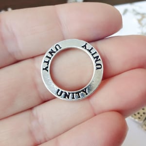 May include: A silver-colored metal charm with the word "UNITY" in black lettering around the outer edge. The charm is circular with a hole in the center. The charm is held in a person's hand.