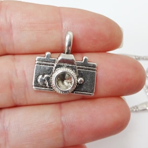 May include: Silver camera charm pendant with a detailed design. The charm has a lens, a shutter button, and a film rewind knob.