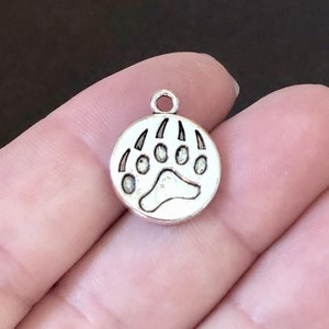May include: Silver charm with a bear paw print design. The charm is round and has a loop at the top for attaching to a necklace or bracelet.
