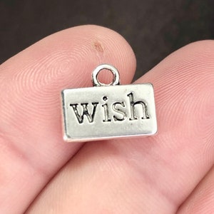 May include: A small, rectangular silver charm with the word "wish" in a bold, serif font. The charm has a loop at the top for attaching to a bracelet or necklace. The charm is a simple, elegant design.