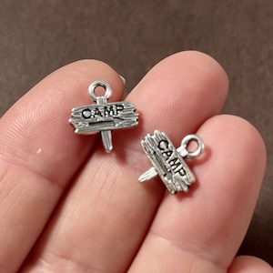 May include: Two silver charm pendants shaped like wooden signs with the word "CAMP" on them. The signs have a rustic, distressed look and are perfect for adding a touch of nature to jewelry.