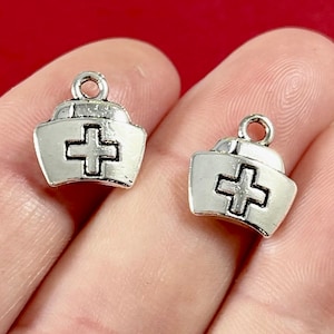 May include: Two silver charm pendants shaped like nurse hats with a white cross in the center. The charms are on a white background.