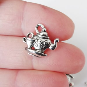 May include: Silver charm in the shape of a teapot and cup. The teapot has a lid and a spout. The cup has a handle. The charm is detailed with swirls and a raised design.