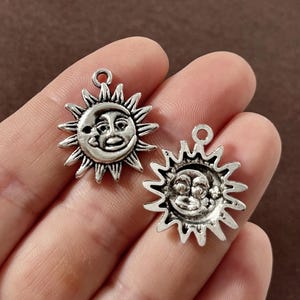 May include: Two silver-toned sun charms with smiling faces and starburst rays. One charm features a crescent moon. The charms are held in a hand, showcasing their intricate details and the metal's aged appearance. These charms are ideal for jewelry making.
