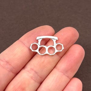 May include: A small, silver-colored knuckle duster with five finger holes. The knuckles are held in a hand against a brown background. The knuckles are a decorative item.